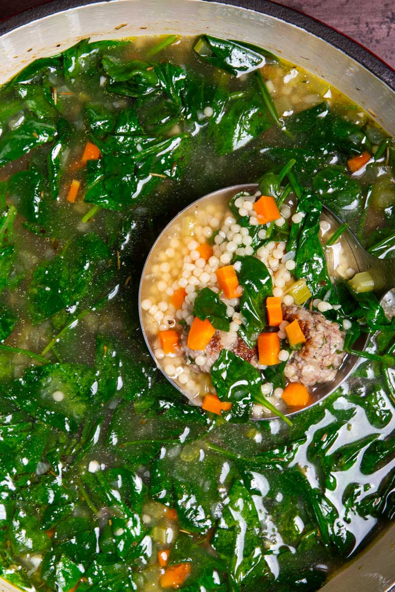 A ladle holding a spoonful of Italian Wedding Soup with seared Italian meatballs, broth, and lots of ancini di pepe pasta, floating over a big pot of steaming soup.