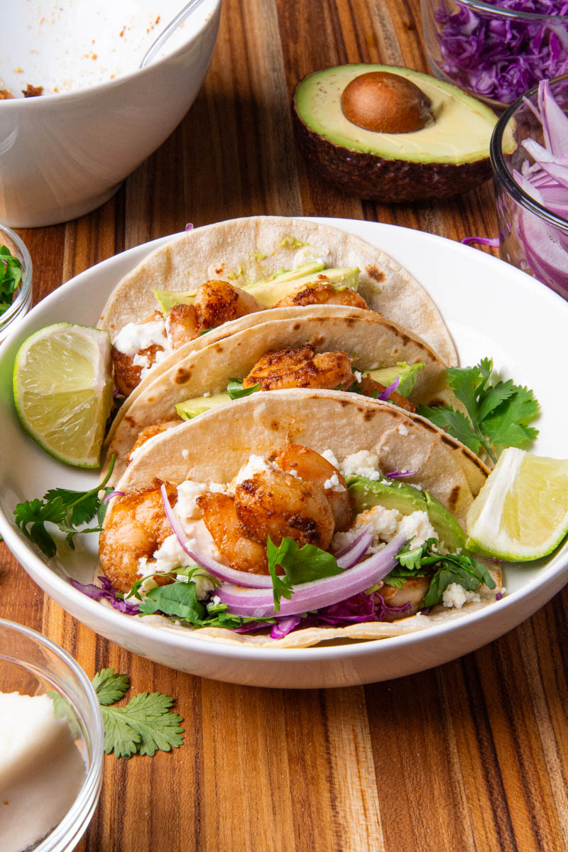 Shrimp tacos in a white bowl, topped with red onion, cotija cheese, avocado, cilantro, red onion, and red cabbage slaw, served with lime wedges, a delicious creamy sauce on a wood cutting board.