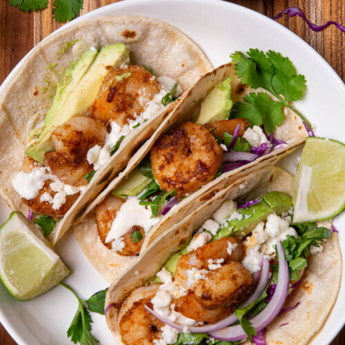 Shrimp tacos in a white bowl, topped with red onion, cotija cheese, avocado, cilantro, red onion, and red cabbage slaw, served with lime wedges, a delicious creamy sauce on a wood cutting board.