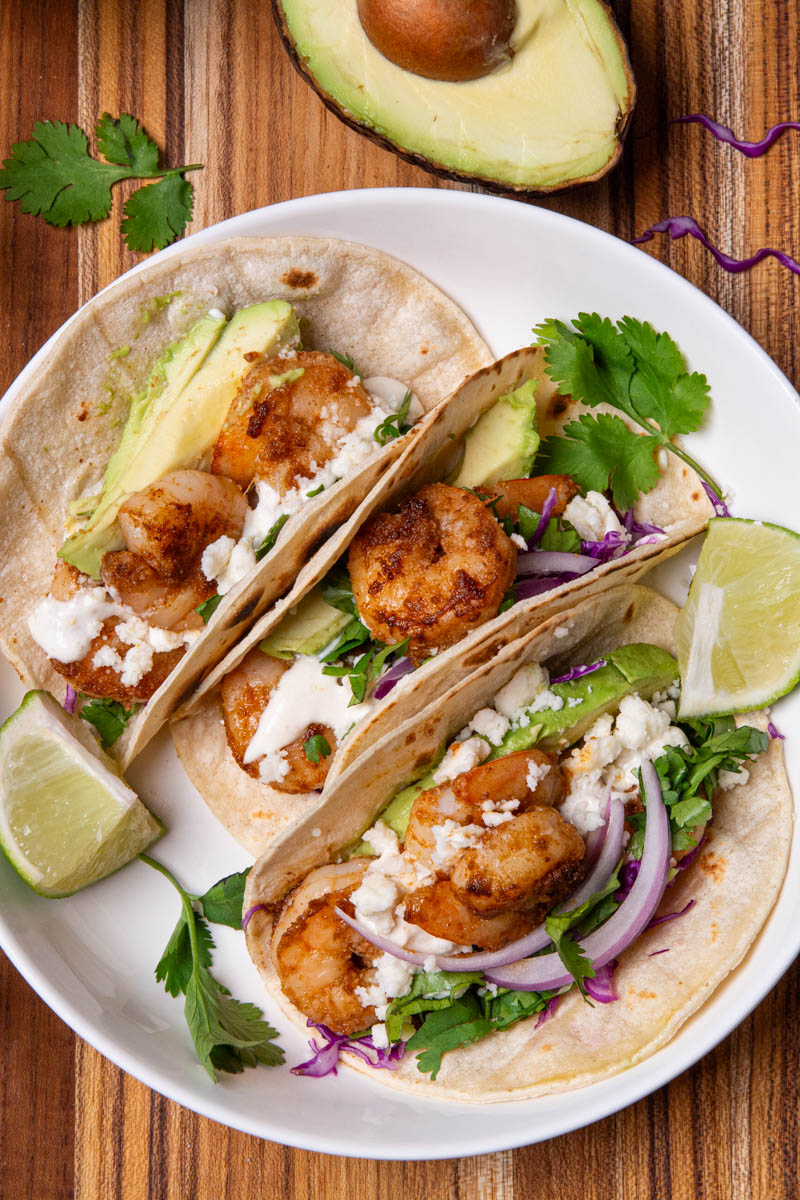 Shrimp tacos in a white bowl, topped with red onion, cotija cheese, avocado, cilantro, red onion, and red cabbage slaw, served with lime wedges, a delicious creamy sauce on a wood cutting board.