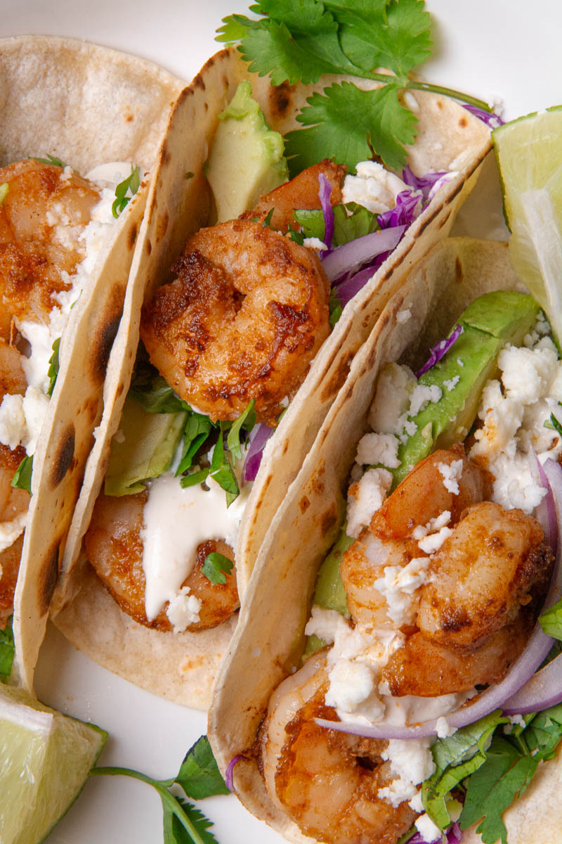 Shrimp tacos in a white bowl, topped with red onion, cotija cheese, avocado, cilantro, red onion, and red cabbage slaw, served with lime wedges, a delicious creamy sauce.