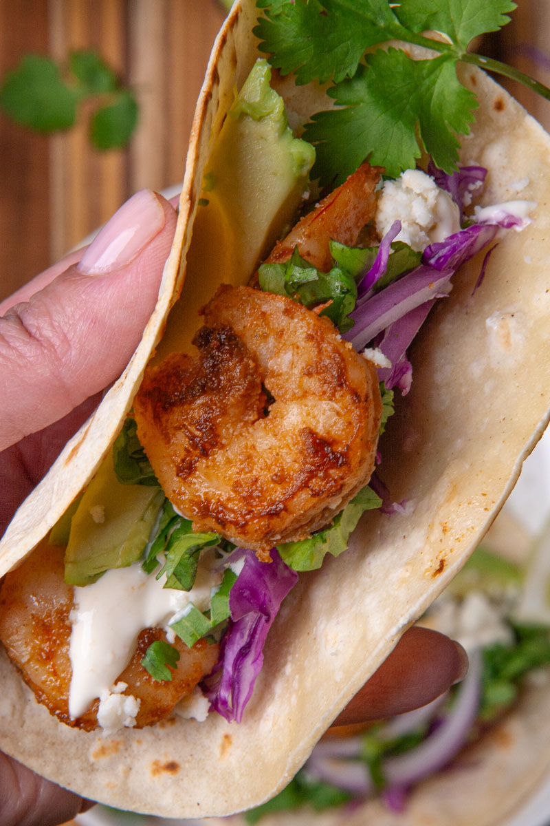 A hand holding a shrimp taco, topped with red onion, cotija cheese, avocado, cilantro, red onion, and red cabbage slaw, served with lime wedges, a delicious creamy sauce.