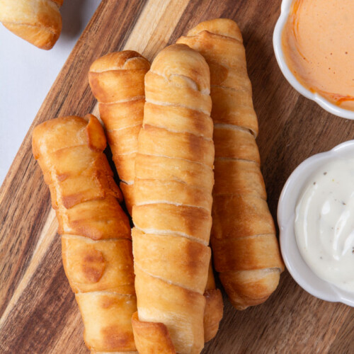 Four golden, crispy Colombian palitos de queso (cheese sticks) or Venezuelan tequeños arranged on a wood cutting board, served with small bowls of ranch and pink sauce for dipping.
