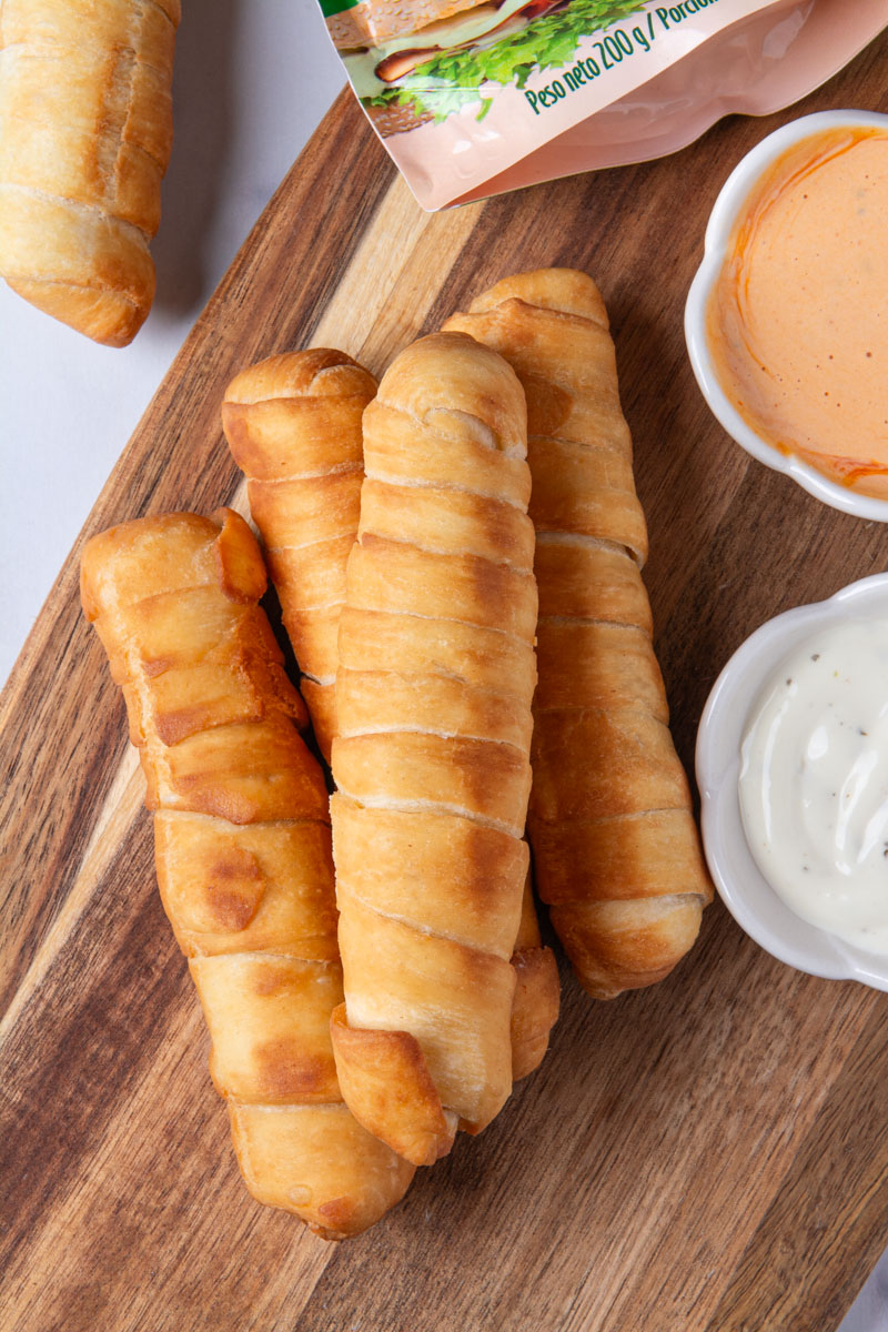 Four golden, crispy Colombian palitos de queso (cheese sticks) or Venezuelan tequeños arranged on a wood cutting board, served with small bowls of ranch and pink sauce for dipping.