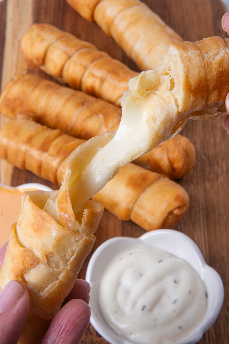 Hands pulling apart a golden, crispy Colombian palito de queso (cheese stick) to reveal a melty mozzarella cheese pull, with more palitos de queso on a wood cutting board and a small bowl of ranch dipping sauce nearby.
