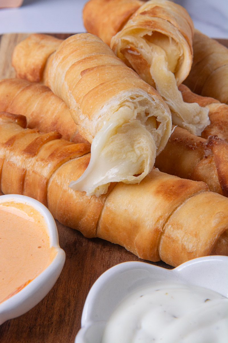 A broken-open Colombian palito de queso (cheese stick) or Venezuelan tequeño showing a melty mozzarella cheese filling, surrounded by more cheese sticks on a wood cutting board with small bowls of pink sauce and ranch for dipping.