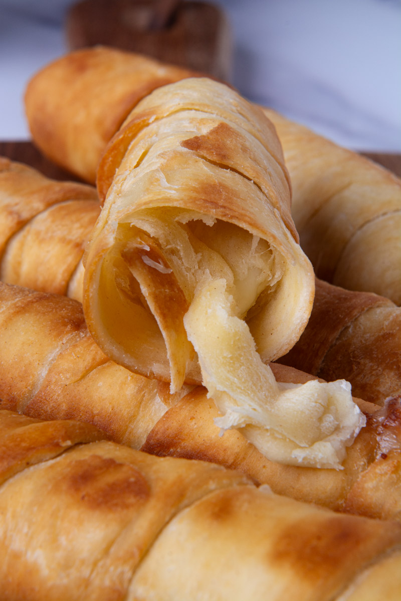 A broken-open Colombian palito de queso (cheese stick) or Venezuelan tequeño showing a melty mozzarella cheese filling, surrounded by more cheese sticks on a wood cutting board with small bowls of pink sauce and ranch for dipping.