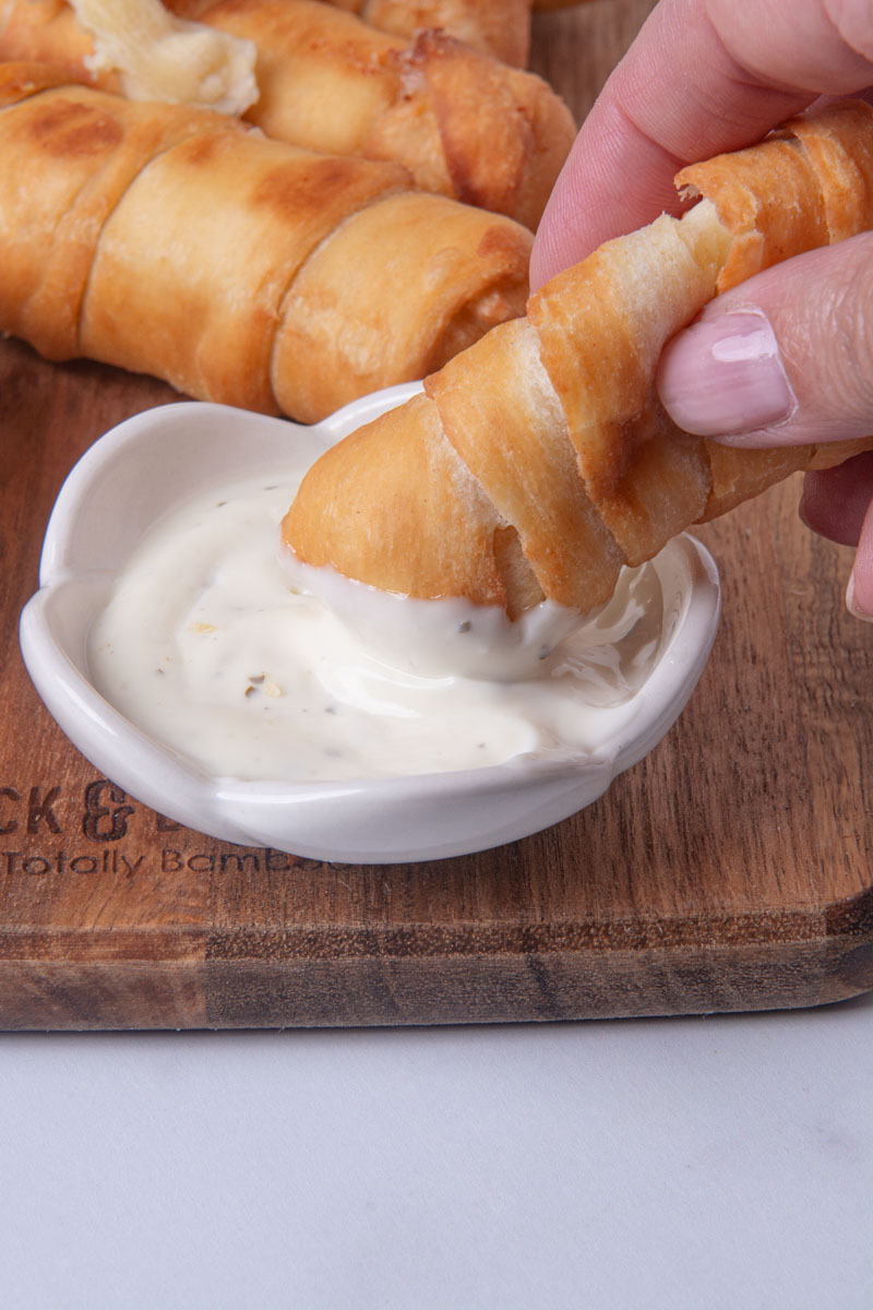 A hand dipping a golden, crispy Colombian palito de queso (cheese stick) or Venezuelan tequeño into a small bowl of ranch sauce on a wood cutting board, with more cheese sticks in the background.