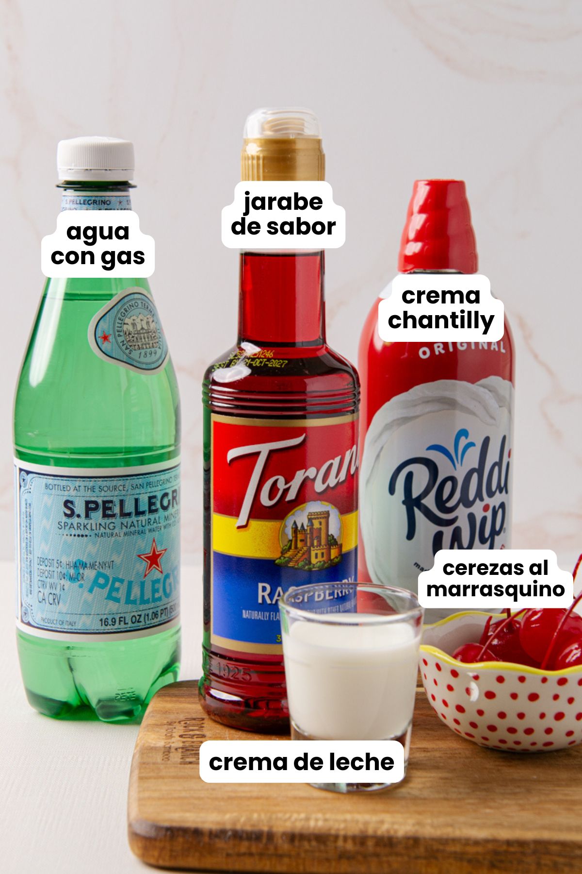 Flat lay of Italian cream soda ingredients on a wood cutting board: a bottle of S.Pellegrino sparkling water, Torani raspberry flavored syrup, Reddi Whip whipped cream, a shot glass of half and half, and a bowl of maraschino cherries, each labeled in Spanish with text overlays.