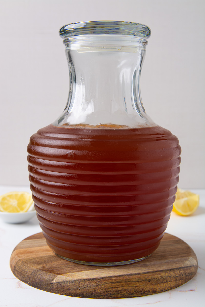 A glass pitcher filled with sweet iced tea on a round wood cutting board, with halved lemons in the background.