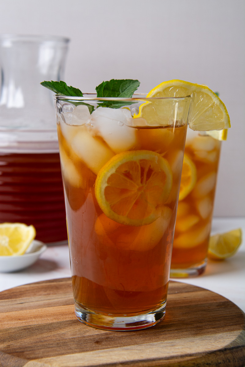 Two tall glasses of sweet iced tea with lemon slices and ice, garnished with fresh mint, on a wood cutting board with a glass pitcher of tea in the background.