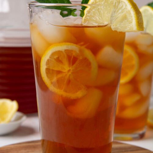 Two tall glasses of sweet iced tea with lemon slices and ice, garnished with fresh mint, on a wood cutting board with a glass pitcher of tea in the background.