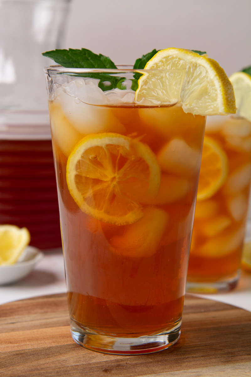 Two tall glasses of sweet iced tea with lemon slices and ice, garnished with fresh mint, on a wood cutting board with a glass pitcher of tea in the background.
