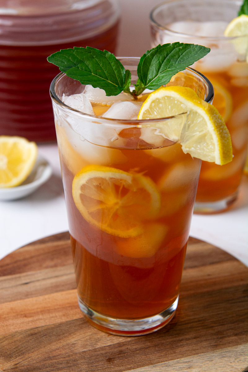 Two tall glasses of sweet iced tea with lemon slices and ice, garnished with fresh mint, on a wood cutting board with a glass pitcher of tea in the background.