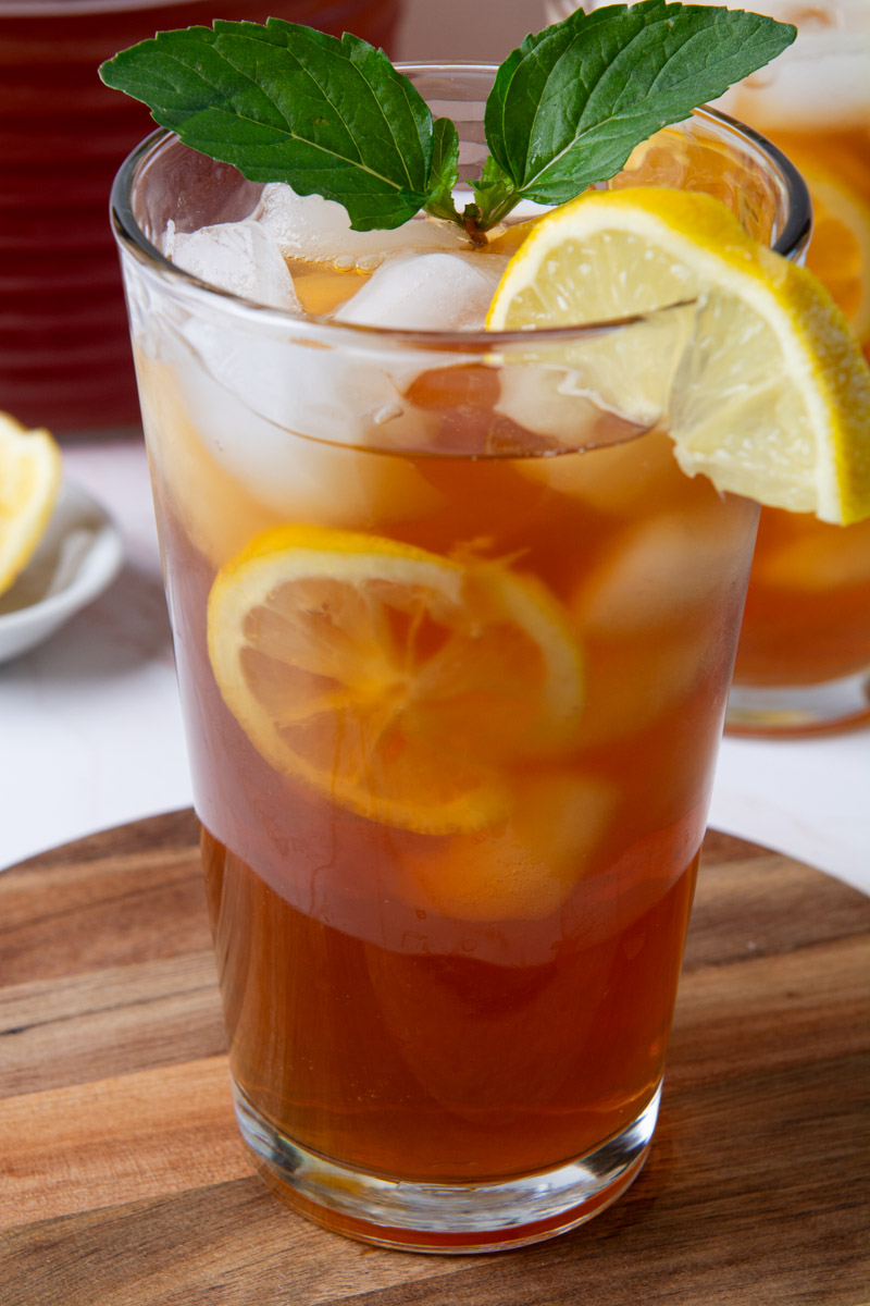 Two tall glasses of sweet iced tea with lemon slices and ice, garnished with fresh mint, on a wood cutting board with a glass pitcher of tea in the background.