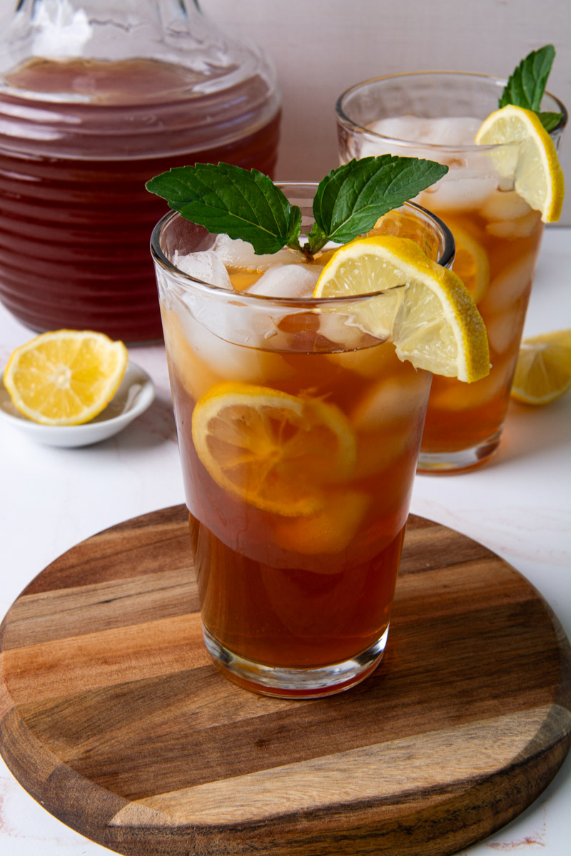 Two tall glasses of sweet iced tea with lemon slices and ice, garnished with fresh mint, on a wood cutting board with a glass pitcher of tea in the background.