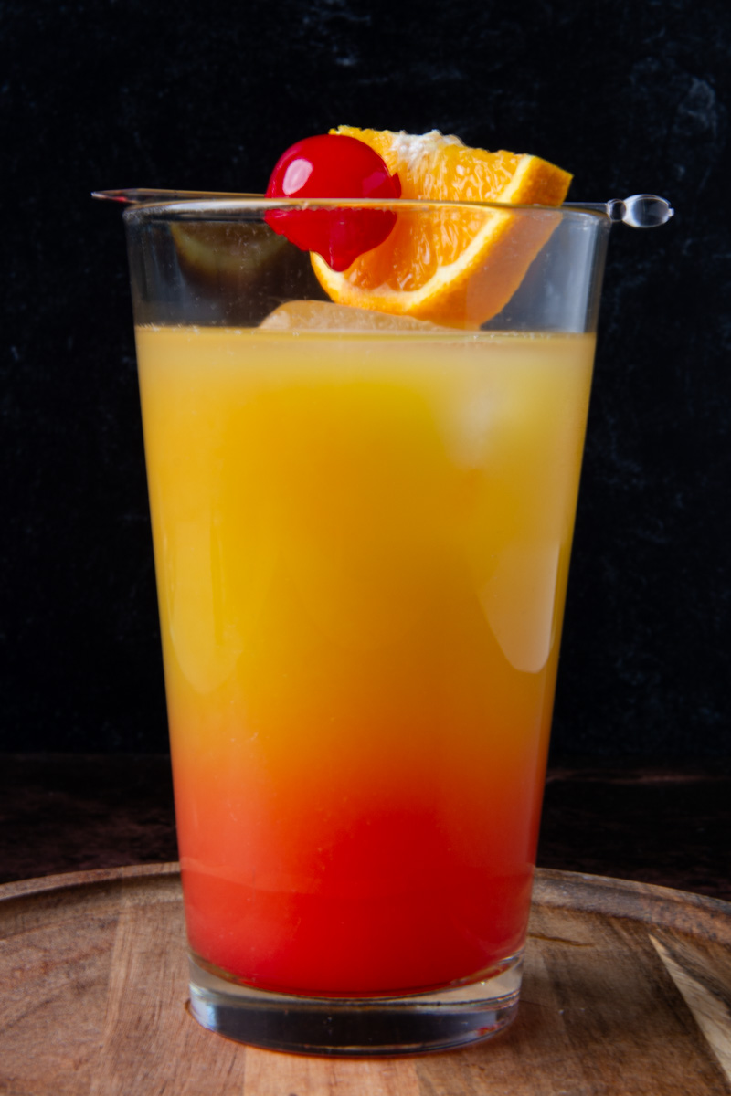 A tequila sunrise cocktail in a tall glass showing a red-to-orange gradient, garnished with a maraschino cherry and orange slice on a cocktail pick, served on a wood surface against a dark background.