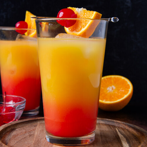 Two tequila sunrise cocktails with a red-to-orange gradient, each garnished with a maraschino cherry and orange slice on a cocktail pick, on a round wood board with a halved orange and a small bowl of cherries nearby.