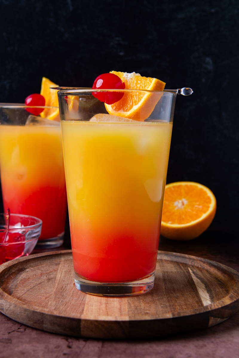 Two tequila sunrise cocktails with a red-to-orange gradient, each garnished with a maraschino cherry and orange slice on a cocktail pick, on a round wood board with a halved orange and a small bowl of cherries nearby.