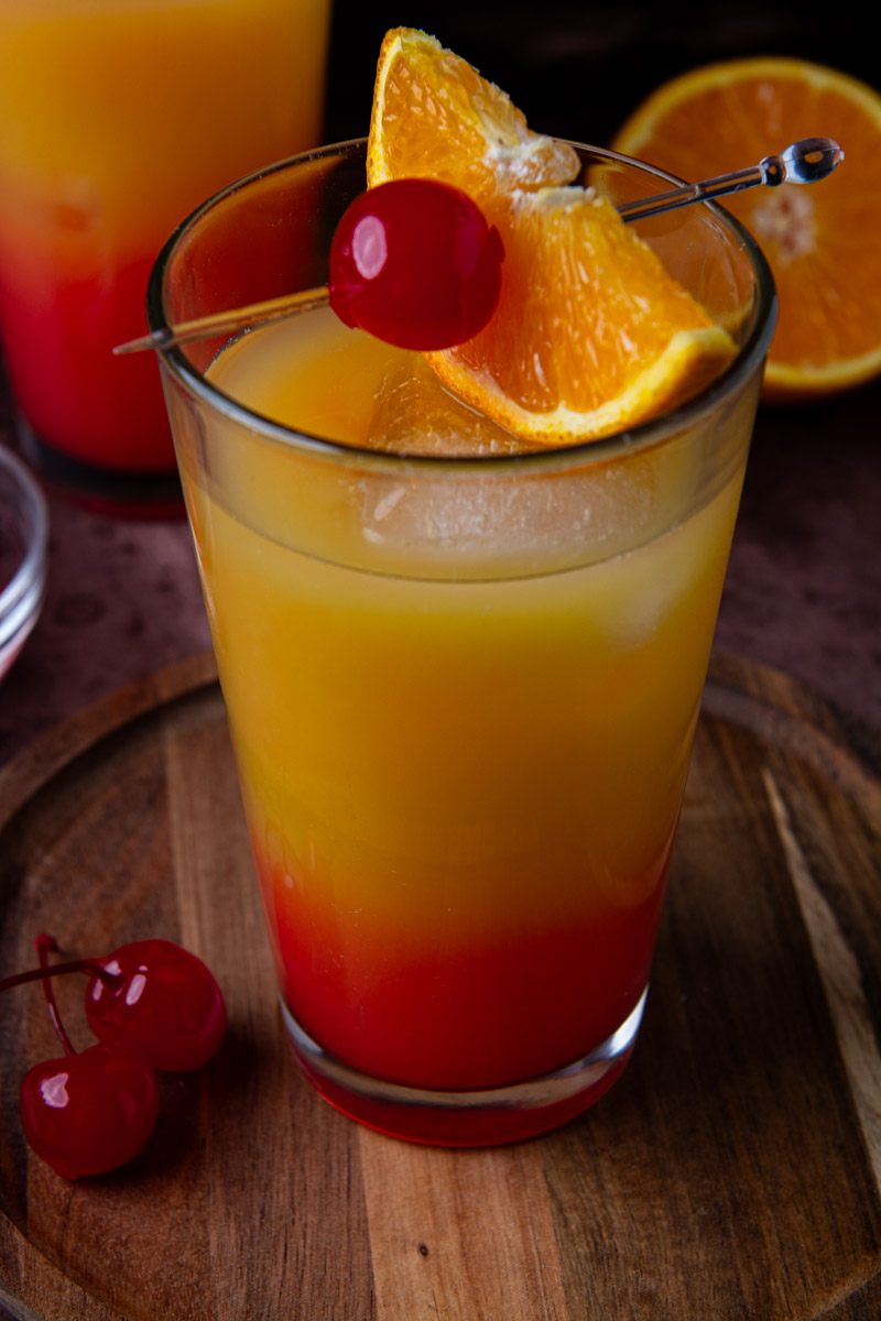 Close-up of a tequila sunrise cocktail with a deep red-to-orange gradient, garnished with a maraschino cherry and orange wedge on a cocktail pick, with loose cherries on the wood board and a second cocktail and halved orange in the background.