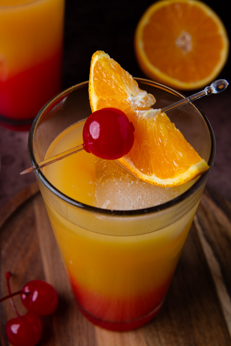Close-up of a tequila sunrise cocktail with a deep red-to-orange gradient, garnished with a maraschino cherry and orange wedge on a cocktail pick, with loose cherries on the wood board and a second cocktail and halved orange in the background.