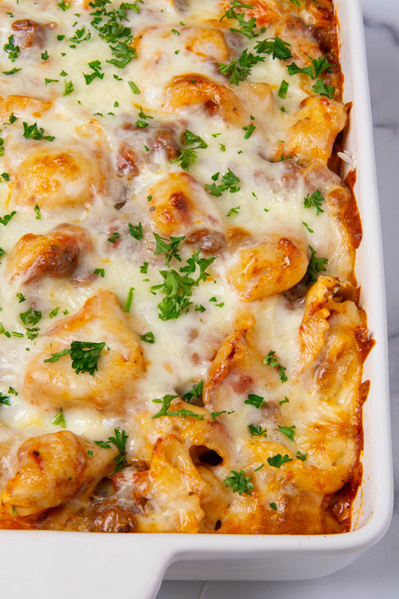 A white baking dish filled with baked cheese tortellini in meat sauce, topped with golden, bubbly melted mozzarella and garnished with fresh chopped parsley.
