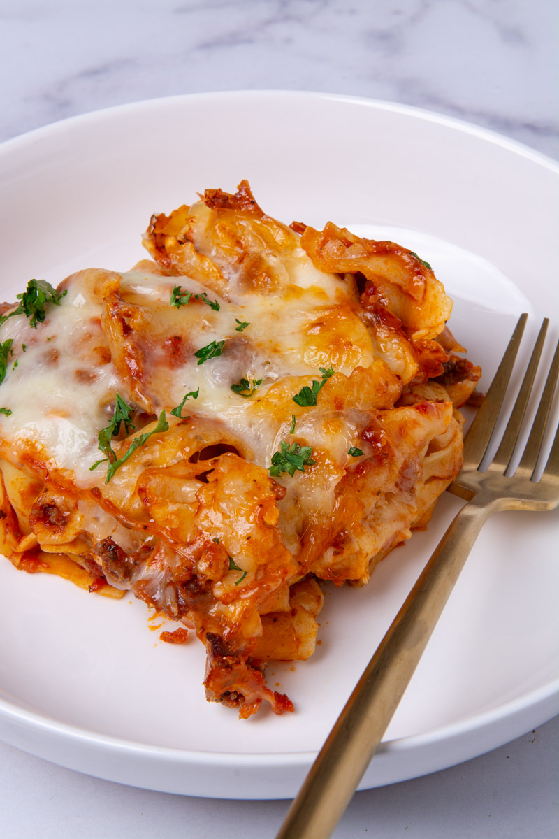A serving of baked cheese tortellini with meat sauce on a white plate, topped with melted mozzarella and fresh parsley, with a gold fork on the side.
