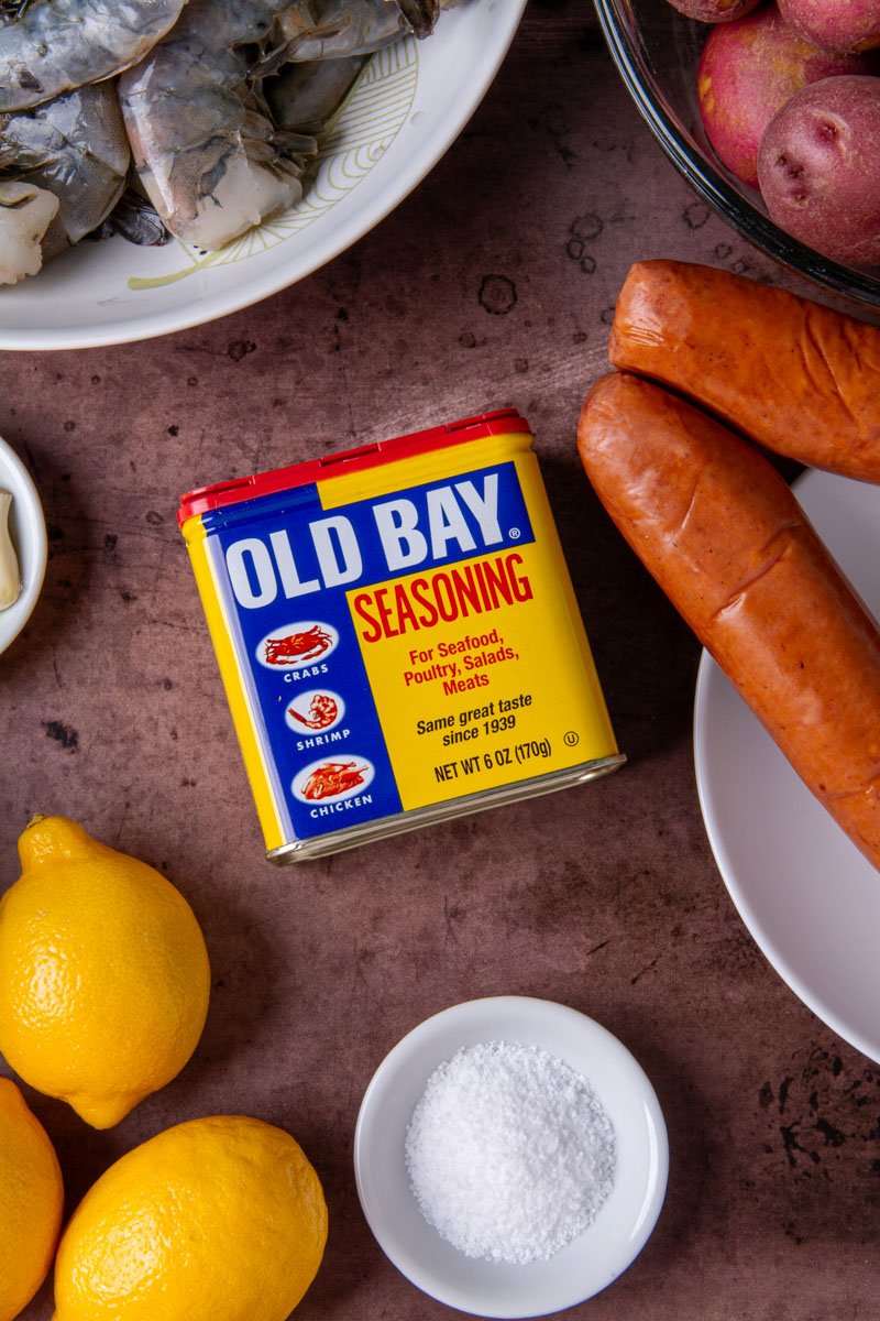 Close-up of a can of Old Bay Seasoning surrounded by shrimp boil ingredients: raw shrimp, smoked sausage, red potatoes, lemons, and a small bowl of salt, on a dark surface.