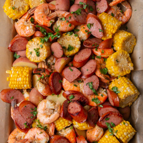 Overhead view of a sheet pan shrimp boil lined with parchment paper, loaded with seasoned shrimp, sliced smoked sausage, corn on the cob, and red potatoes, garnished with fresh chopped cilantro.