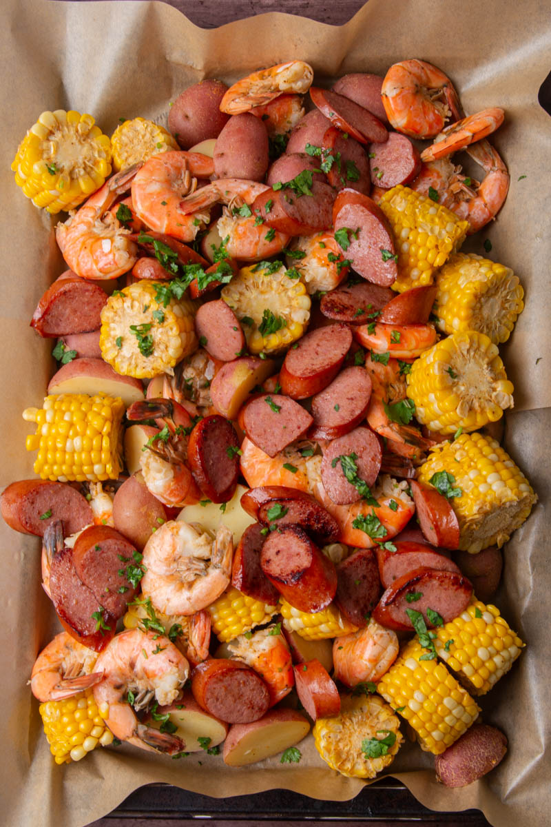 Overhead view of a sheet pan shrimp boil lined with parchment paper, loaded with seasoned shrimp, sliced smoked sausage, corn on the cob, and red potatoes, garnished with fresh chopped cilantro.