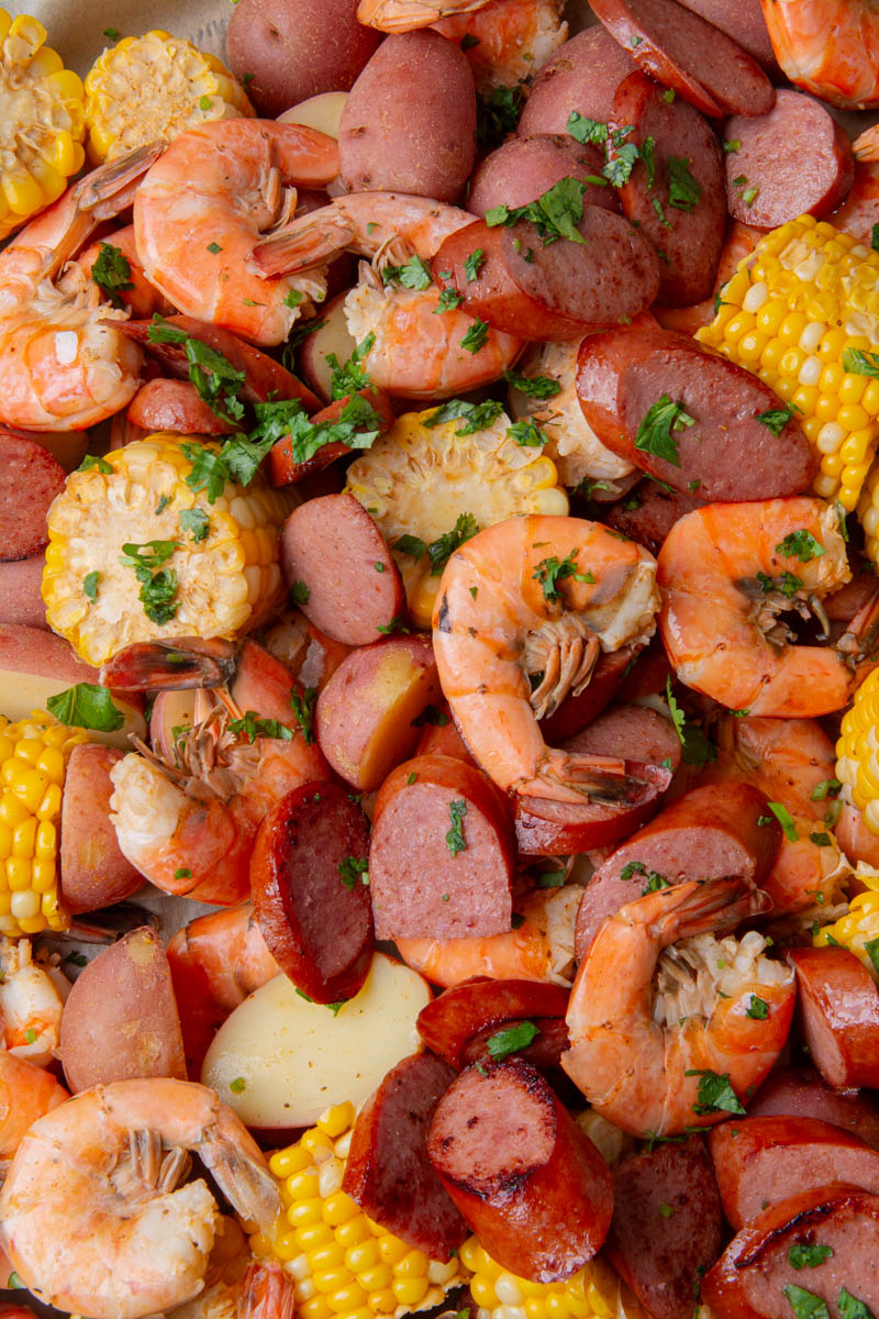 Close-up of a sheet pan shrimp boil with seasoned shrimp, sliced smoked sausage, corn on the cob, and red potatoes, all garnished with fresh chopped cilantro.