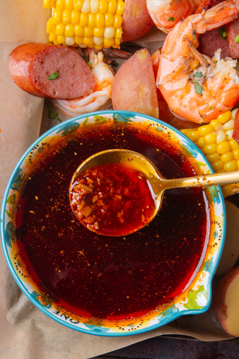 Close-up of a gold spoon lifting garlic-spiced butter sauce from a decorative bowl, with a sheet pan seafood boil of shrimp, smoked sausage, corn, and red potatoes in the background.