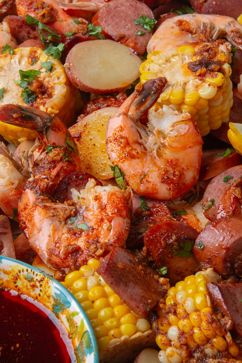 Close-up of a sheet pan shrimp boil coated in spiced butter sauce, with juicy shrimp, sliced smoked sausage, corn on the cob, and red potatoes, garnished with fresh chopped cilantro.