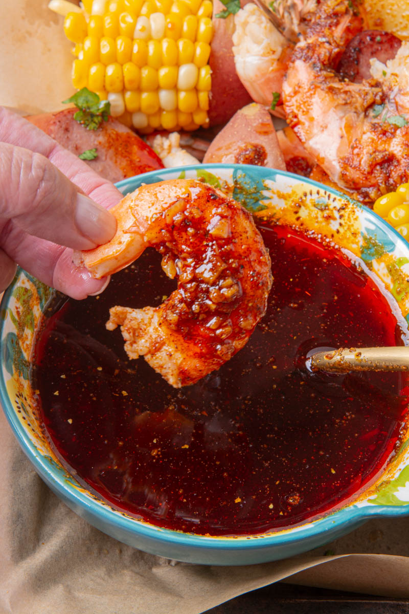 A hand dipping a seasoned shrimp into a bowl of spiced garlic butter sauce, with a sheet pan seafood boil of corn, shrimp, sausage, and potatoes in the background.