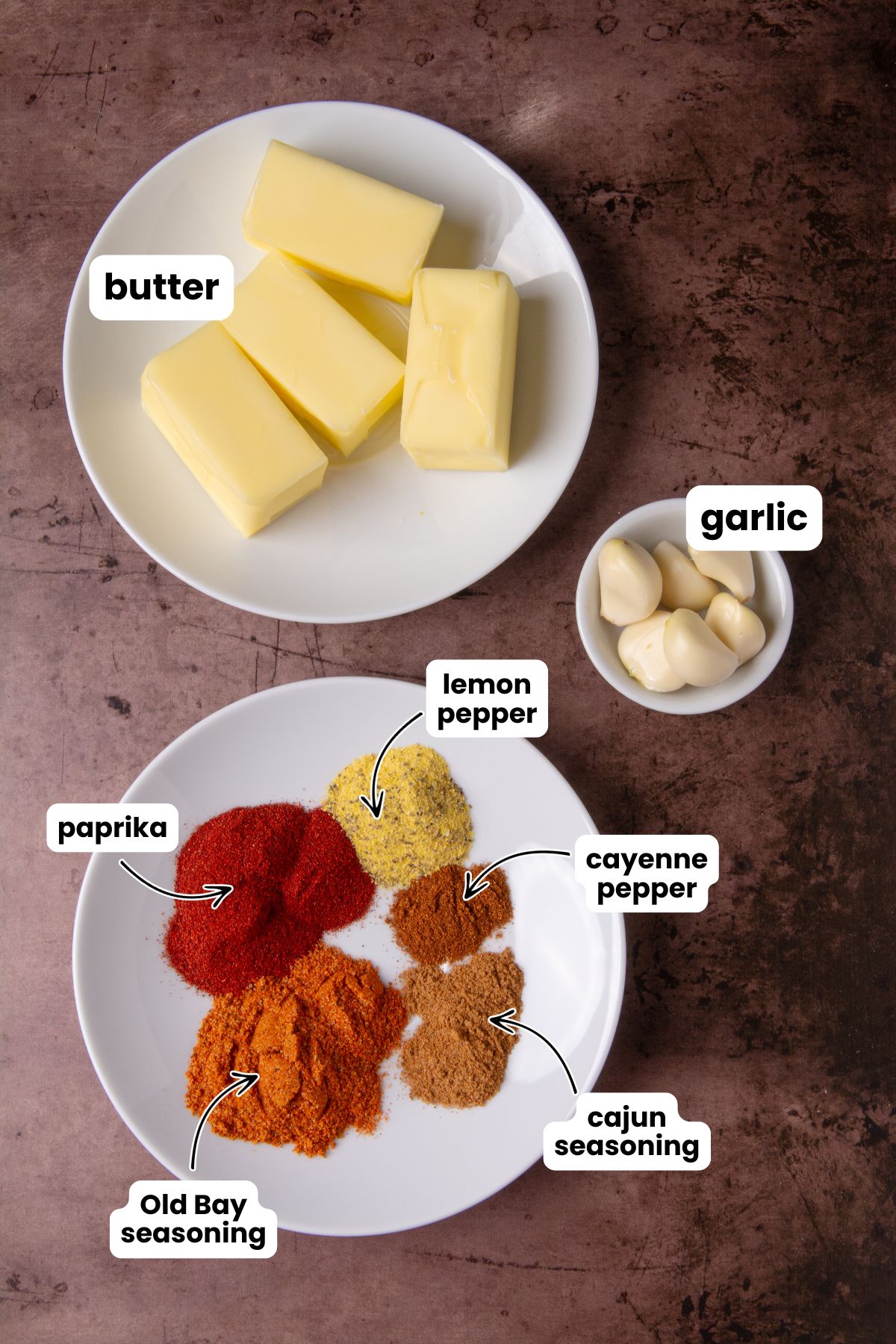 Overhead flat lay of shrimp boil butter sauce ingredients on a dark surface: a plate with butter sticks, a bowl of garlic cloves, and a plate with paprika, lemon pepper, cayenne pepper, Old Bay seasoning, and Cajun seasoning, each labeled with text overlays.