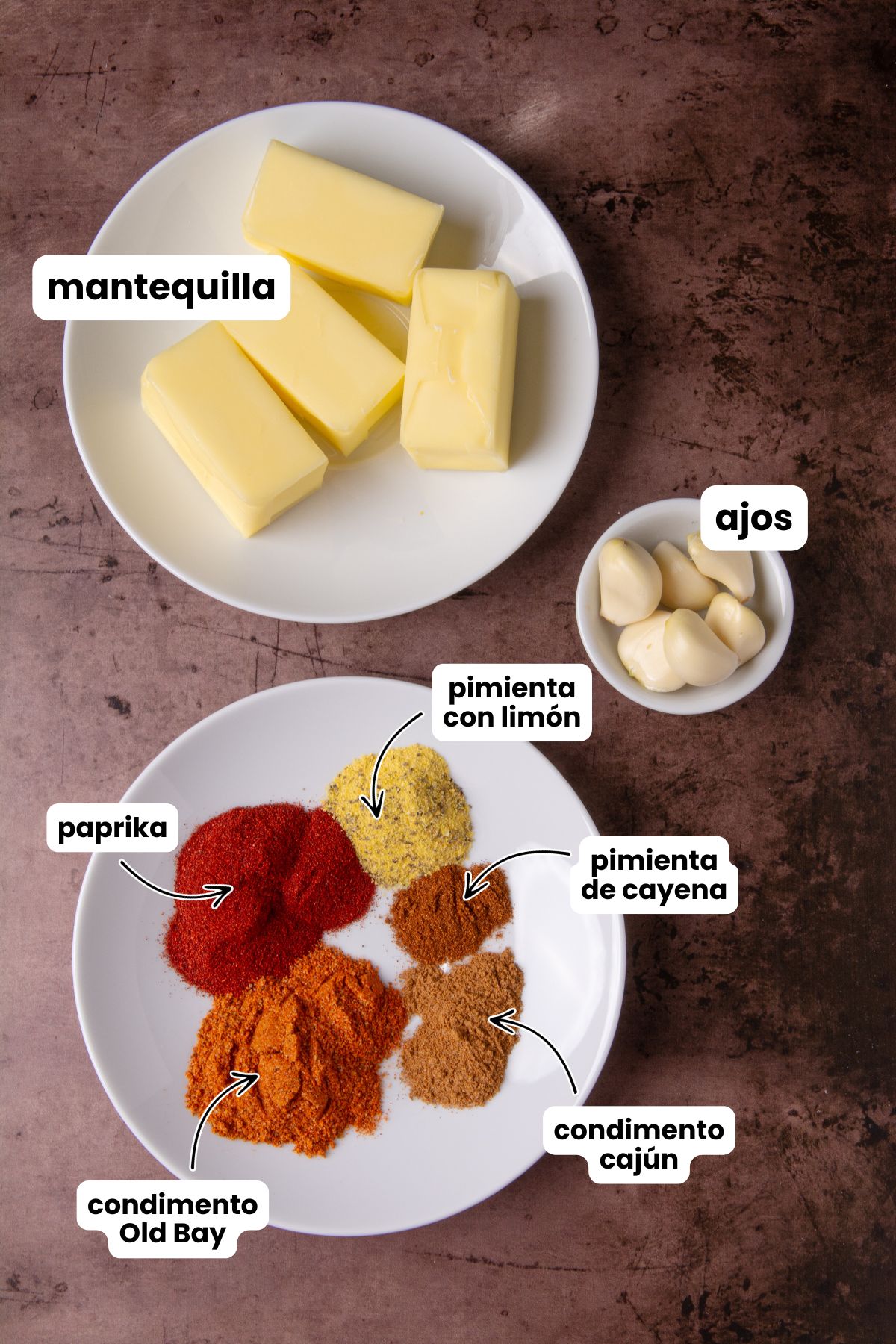Overhead flat lay of shrimp boil butter sauce ingredients on a dark surface: a plate with butter sticks, a bowl of garlic cloves, and a plate with paprika, lemon pepper, cayenne pepper, Old Bay seasoning, and Cajun seasoning, each labeled with Spanish text overlays.