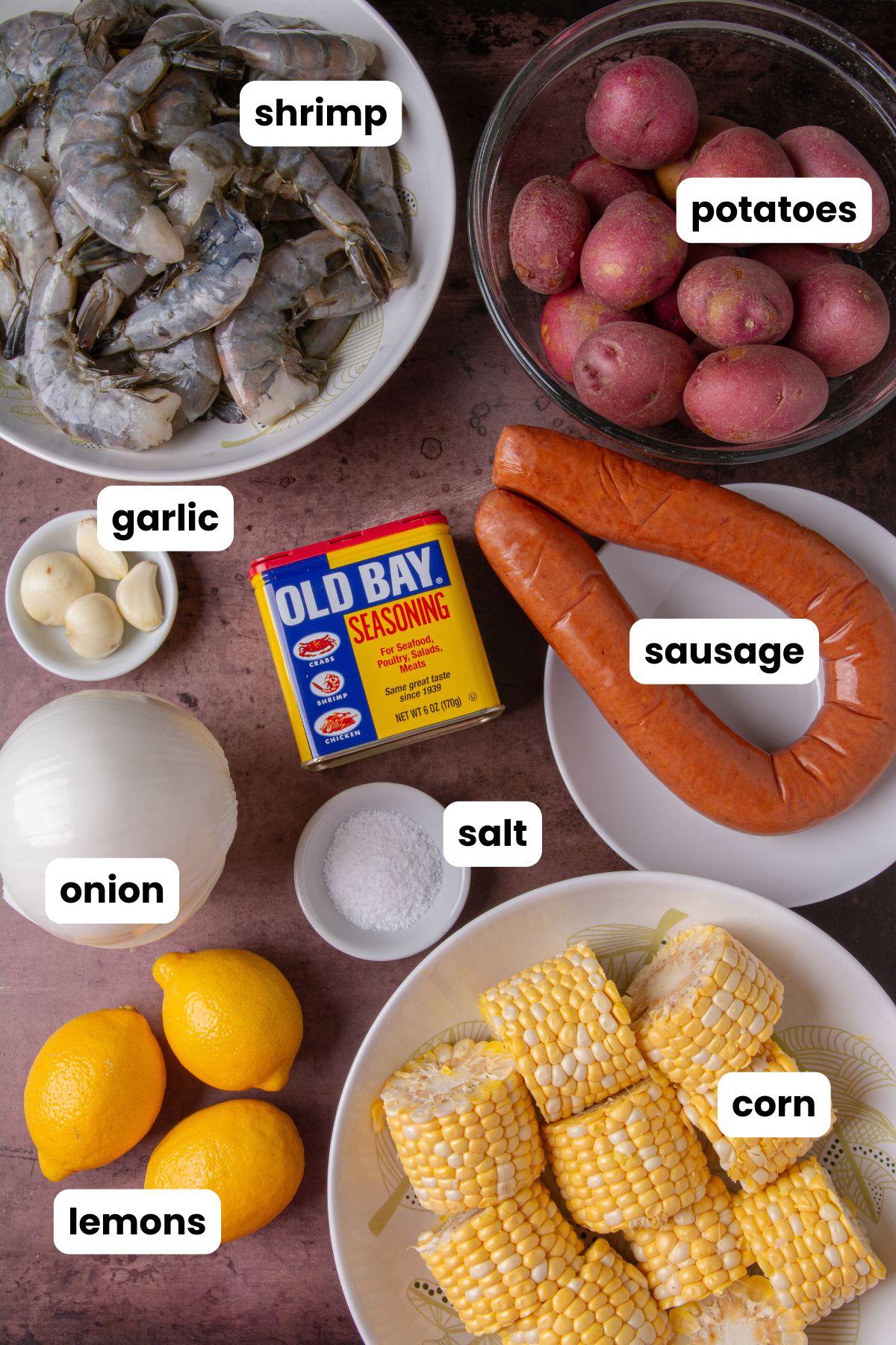 Overhead flat lay of shrimp boil ingredients on a dark surface: raw shrimp, red potatoes, smoked sausage, garlic cloves, a can of Old Bay seasoning, a white onion, salt, lemons, and corn on the cob, each labeled with text overlays.