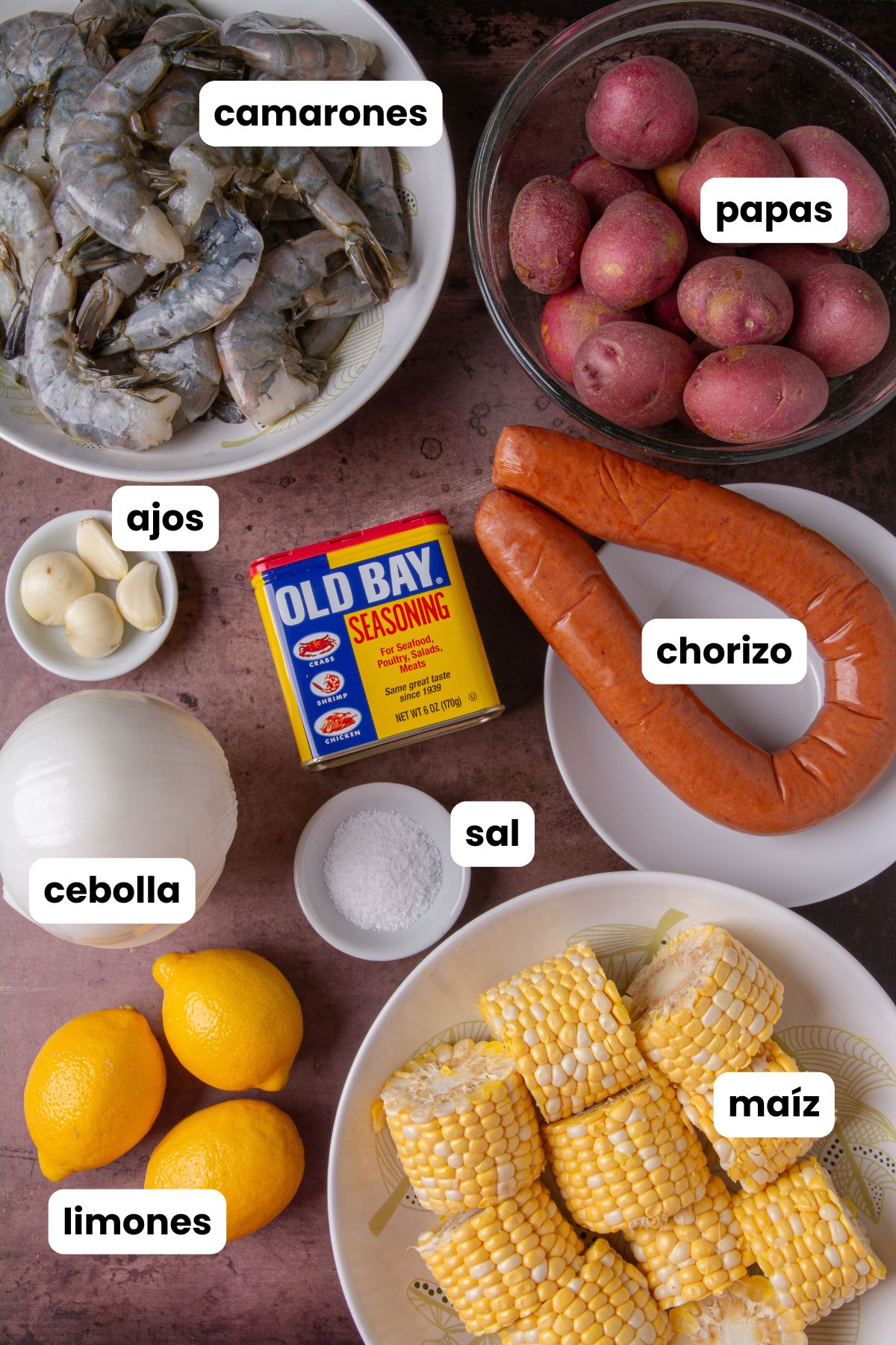 Overhead flat lay of shrimp boil ingredients on a dark surface: raw shrimp, red potatoes, smoked sausage, garlic cloves, a can of Old Bay seasoning, a white onion, salt, lemons, and corn on the cob, each labeled with Spanish text overlays.