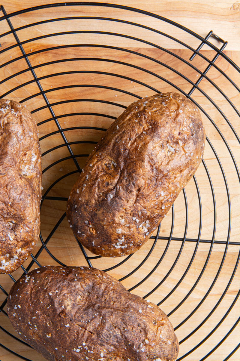 Overhead view of three air fryer baked potatoes with crispy, golden-brown skin sprinkled with coarse salt, resting on a round black wire cooling rack over a wood surface.