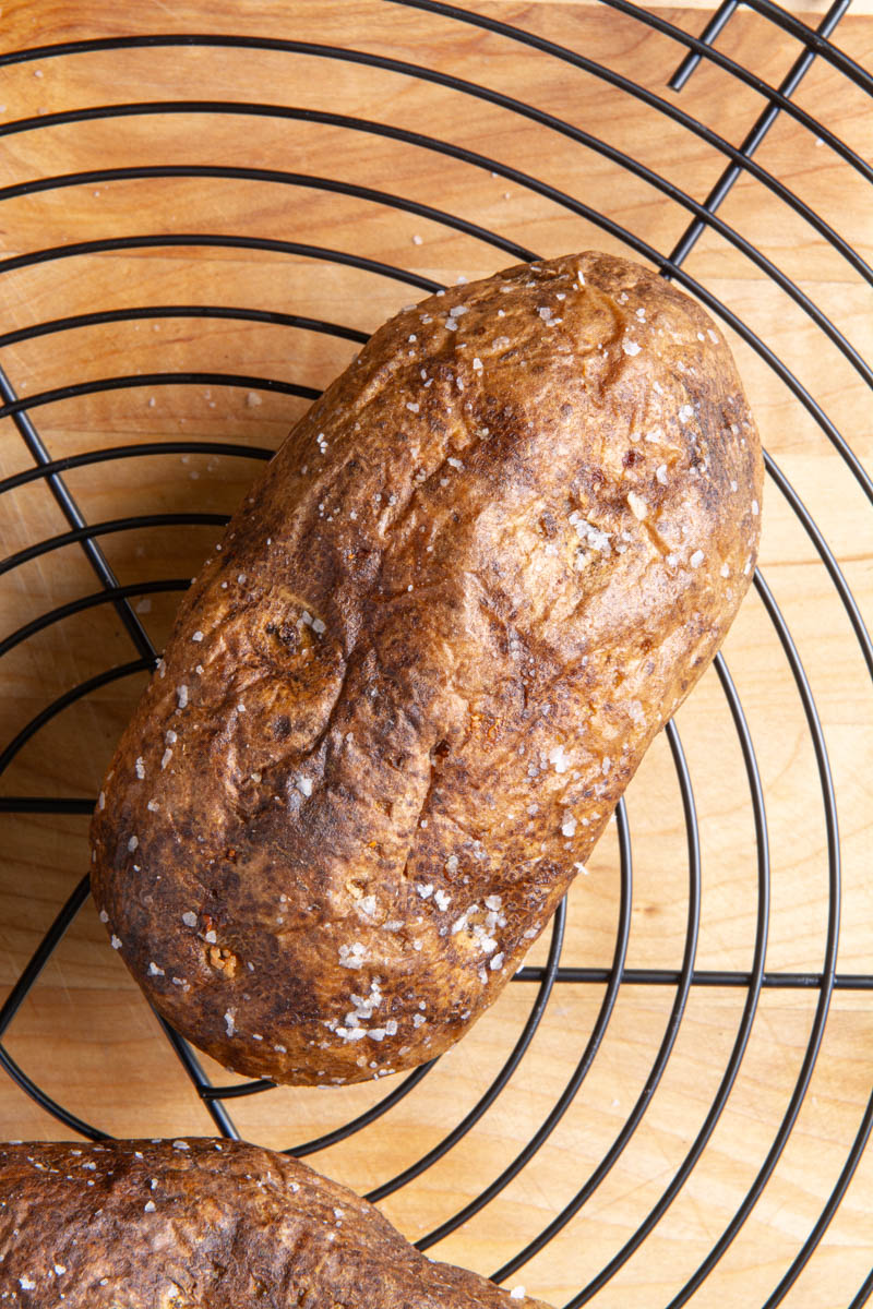 Close-up of an air fryer baked potato with crispy, golden-brown skin sprinkled with coarse salt, on a round black wire rack over a wood surface.