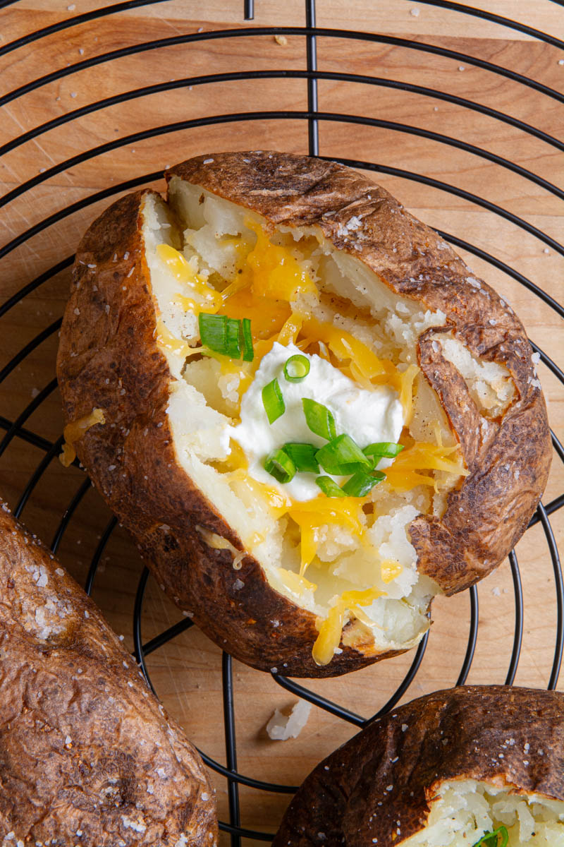 Overhead view of an air fryer baked potato split open and loaded with melted cheddar cheese, sour cream, and sliced green onions, on a round black wire rack with more potatoes in the background.