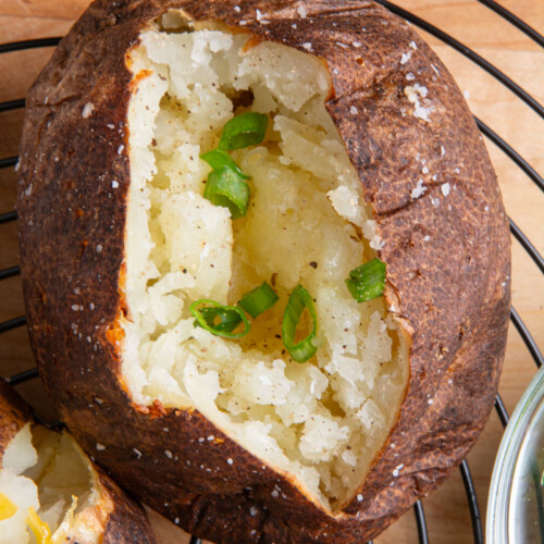 Overhead close-up of an air fryer baked potato split open to reveal a fluffy interior, topped with sliced green onions, on a black wire rack with more potatoes and toppings in the background.