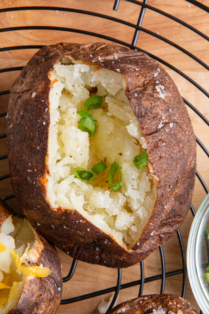 Overhead close-up of an air fryer baked potato split open to reveal a fluffy interior, topped with sliced green onions, on a black wire rack with more potatoes and toppings in the background.