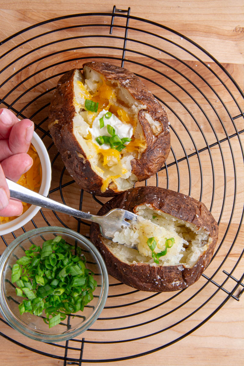 Overhead view of two air fryer baked potatoes on a black wire rack, one loaded with melted cheddar, sour cream, and green onions, the other being fluffed with a fork with small bowls of shredded cheese and chopped green onions nearby.