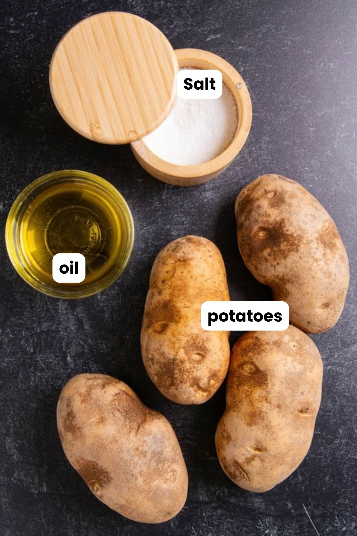 Overhead flat lay of air fryer baked potato ingredients on a dark surface: four russet potatoes, a small bowl of oil, and a wooden salt cellar, each labeled with text overlays.