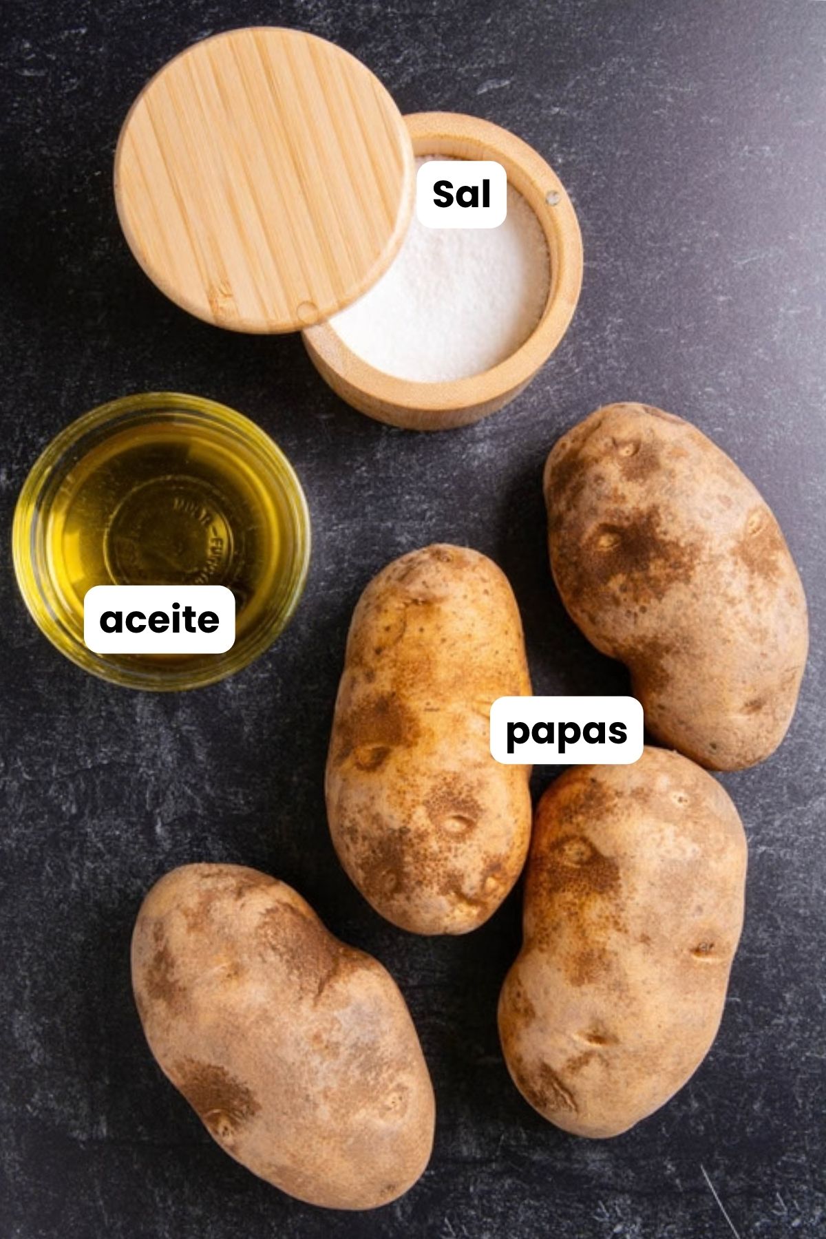 Overhead flat lay of air fryer baked potato ingredients on a dark surface: four russet potatoes, a small bowl of oil, and a wooden salt cellar, each labeled with Spanish text overlays.
