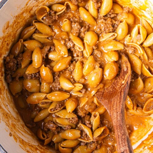 Overhead view of homemade hamburger helper cooking in a red Dutch oven, with pasta shells and ground beef simmering in a rich, seasoned sauce, stirred with a wooden spoon.