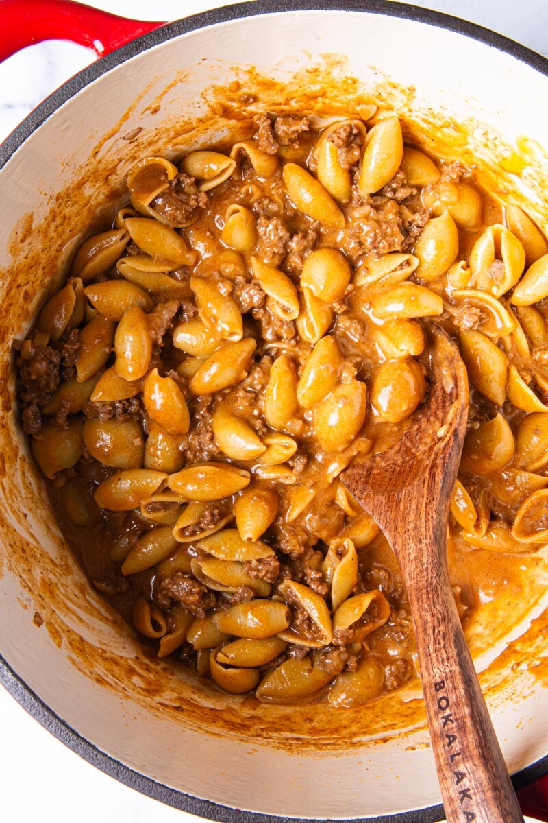 Overhead view of homemade hamburger helper cooking in a red Dutch oven, with pasta shells and ground beef simmering in a rich, seasoned sauce, stirred with a wooden spoon.