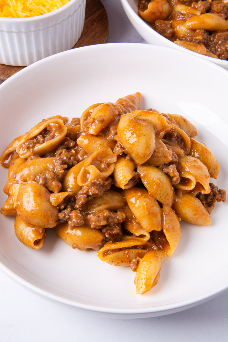 A white bowl of homemade hamburger helper with pasta shells and ground beef in a rich, seasoned sauce, with a bowl of shredded cheddar cheese and a second serving in the background.
