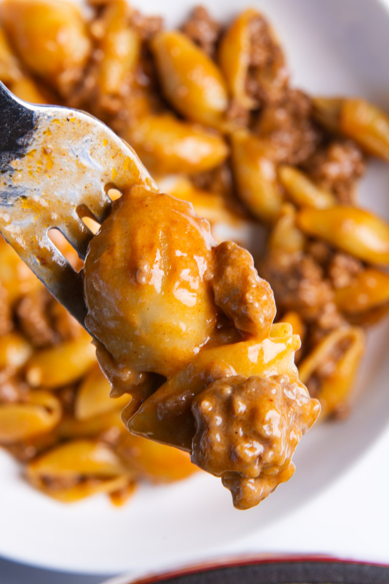 A fork holding a saucy pasta shell and ground beef bite from a bowl of homemade hamburger helper, with the rest of the dish blurred in the background.